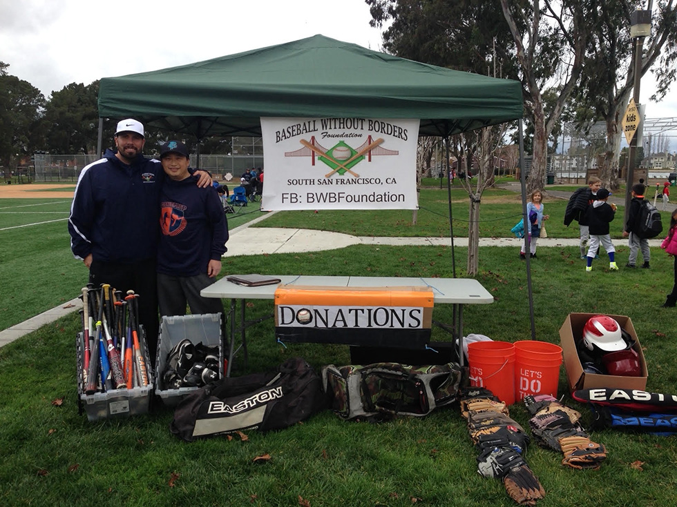 Two volunteers stand beside a donations table and 'Baseball Without Borders Foundation' banner at a South San Francisco baseball field.