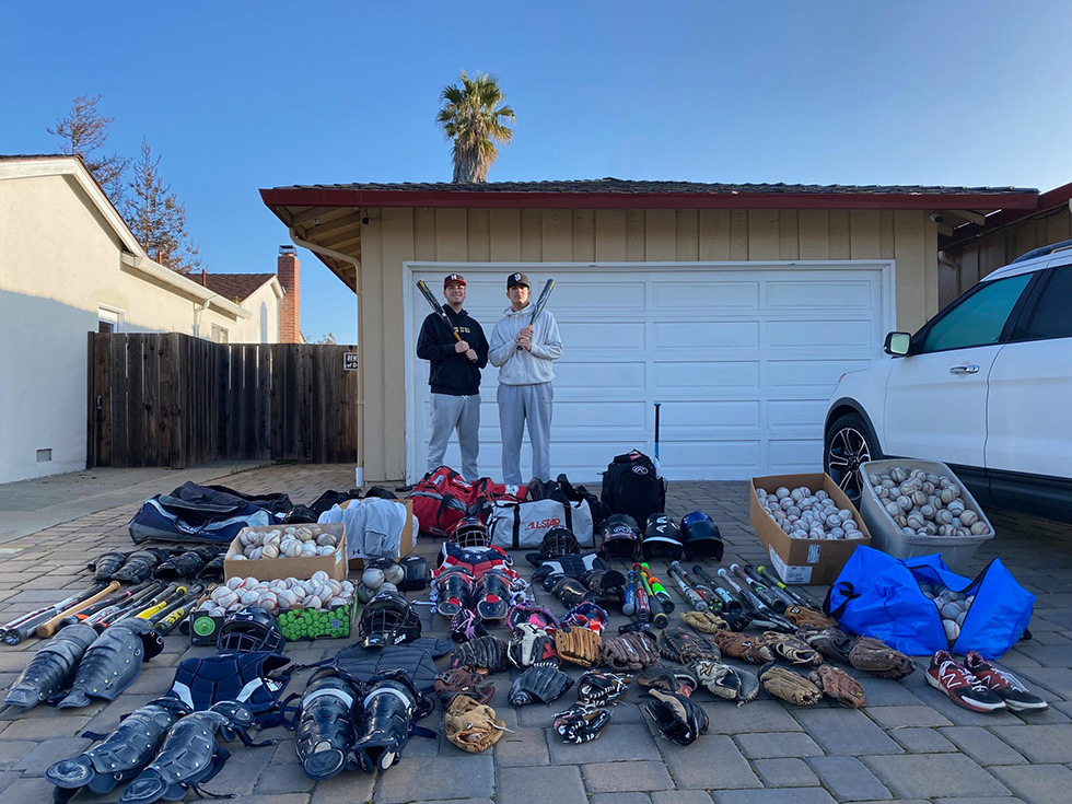 Two volunteers pose with baseball bats beside boxes of donated gloves, balls, and gear for Baseball Without Borders Foundation.