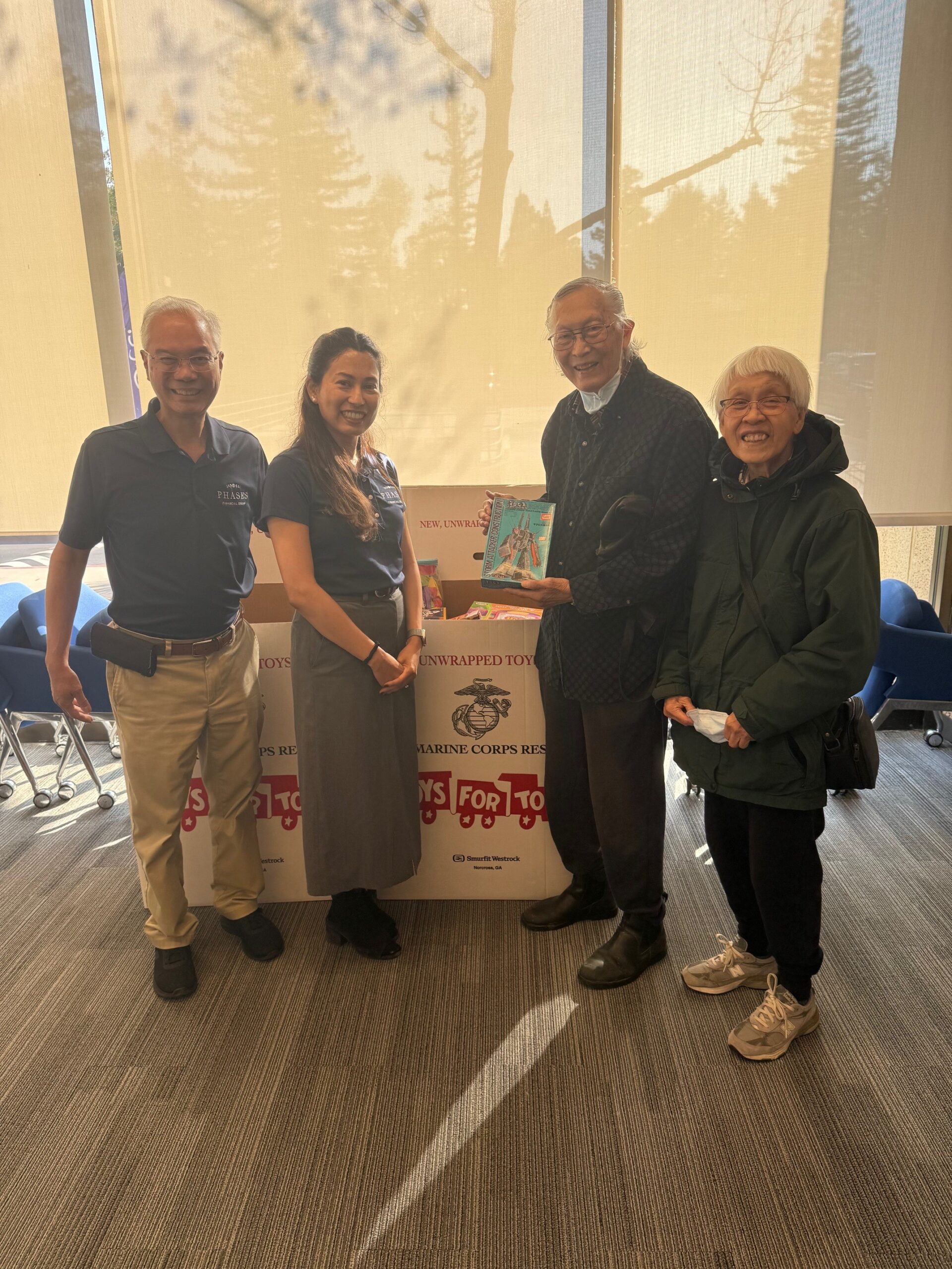 John and Judy Jang donate toys to Phases Financial Group's Toys for Tots collection box, joined by two team members.
